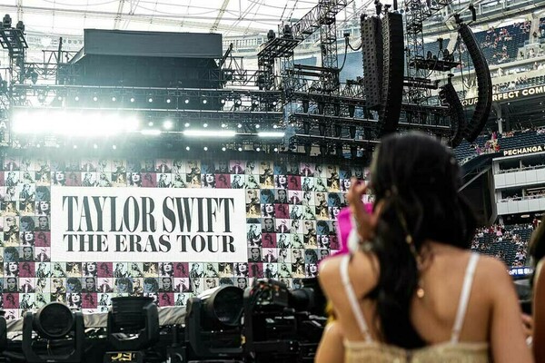 Fans stand in a stadium with a huge banner in the background that reads "Taylor Swift: The Eras Tour"