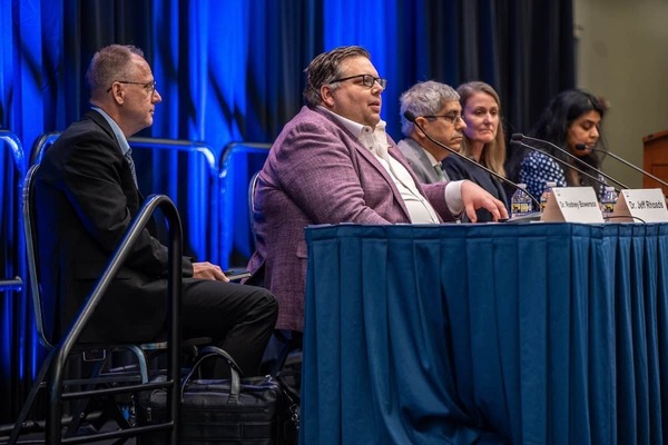 A panel of speakers sit behind a blue table. The panel includes Jeff Rhoads, Vice President for Research.