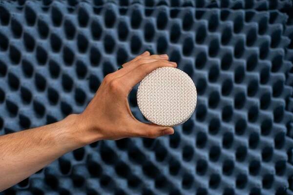 A hand holds a small, circular, white object with a textured, woven pattern. The backdrop is a dark blue surface with a repeating, geometric pattern of concave shapes.