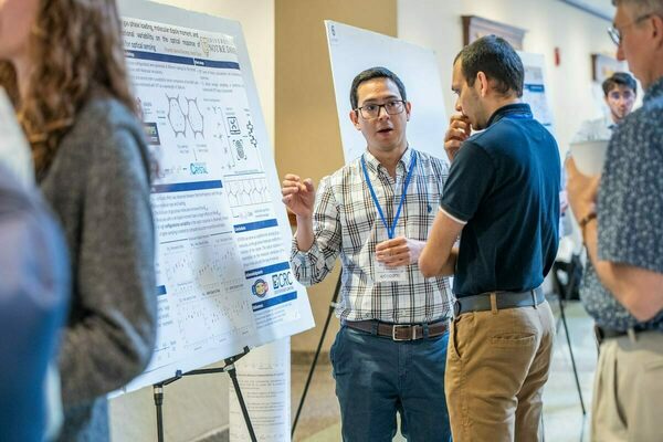 A man in a plaid shirt gestures while explaining his research poster to another man at a University of Notre Dame event.  A third person looks on.  The poster includes scientific charts, diagrams, and text.