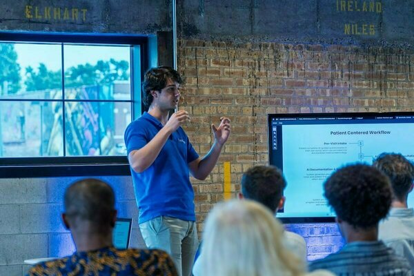 A presenter in a blue shirt gestures while speaking to a small audience in front of a screen displaying "Patient Centered Workflow".
