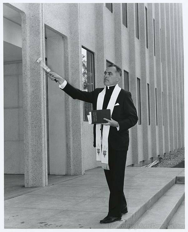 A priest in a black suit, Roman collar, and an embroidered white stole holds a book and raises a holy water sprinkler, blessing the area outside a modern grey building.
