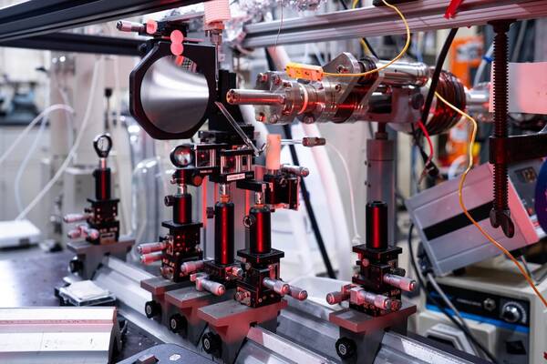 Intricate scientific experiment setup in a lab, featuring metal optical components, precision mounts, and lenses on an optical bench. A large silver funnel and a central bolted cylinder are visible, with red light illuminating several elements and yellow wires connecting the apparatus.