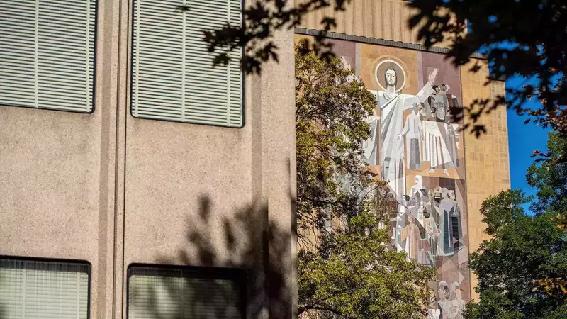 The "Word of Life" mural on the Hesburgh Library features Jesus with raised arms and surrounding figures in mosaic tiles, viewed partially through green-leafed trees.