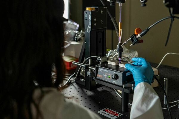 A researcher in a white lab coat and blue glove operates scientific equipment, emitting a glowing purple plasma jet, on a black optical breadboard. A "Danger High Voltage" sticker is visible.