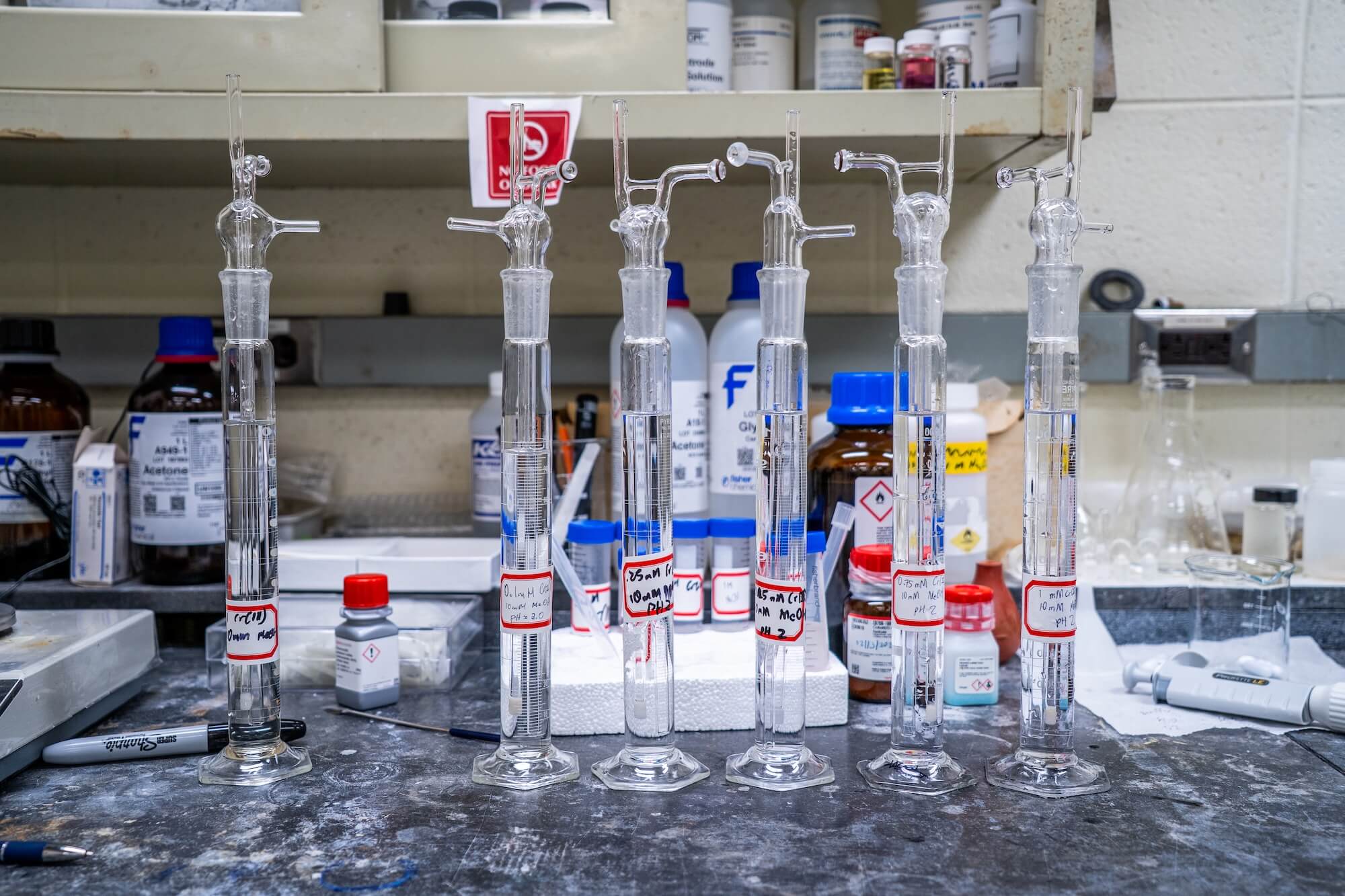 Six clear glass lab instruments, filled with clear liquid and marked with red-bordered labels, stand on a dark lab bench among chemical bottles and lab equipment.