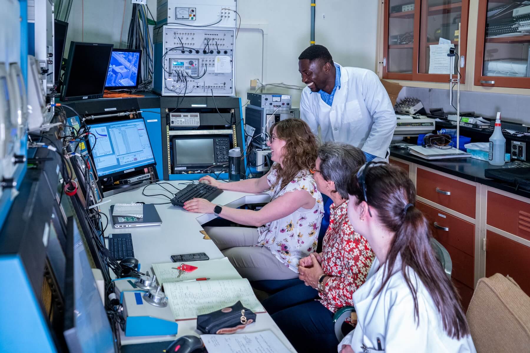 A smiling man in a lab coat looks at three women gathered around a computer workstation in a research lab. One woman types on a keyboard while others observe the screens displaying data.
