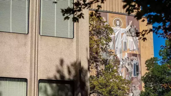 The Word of Life mural on the Hesburgh Library features a mosaic of Jesus with arms raised, surrounded by numerous saints and scholars. Green tree branches partially frame the colorful mosaic and tan building under a blue sky.