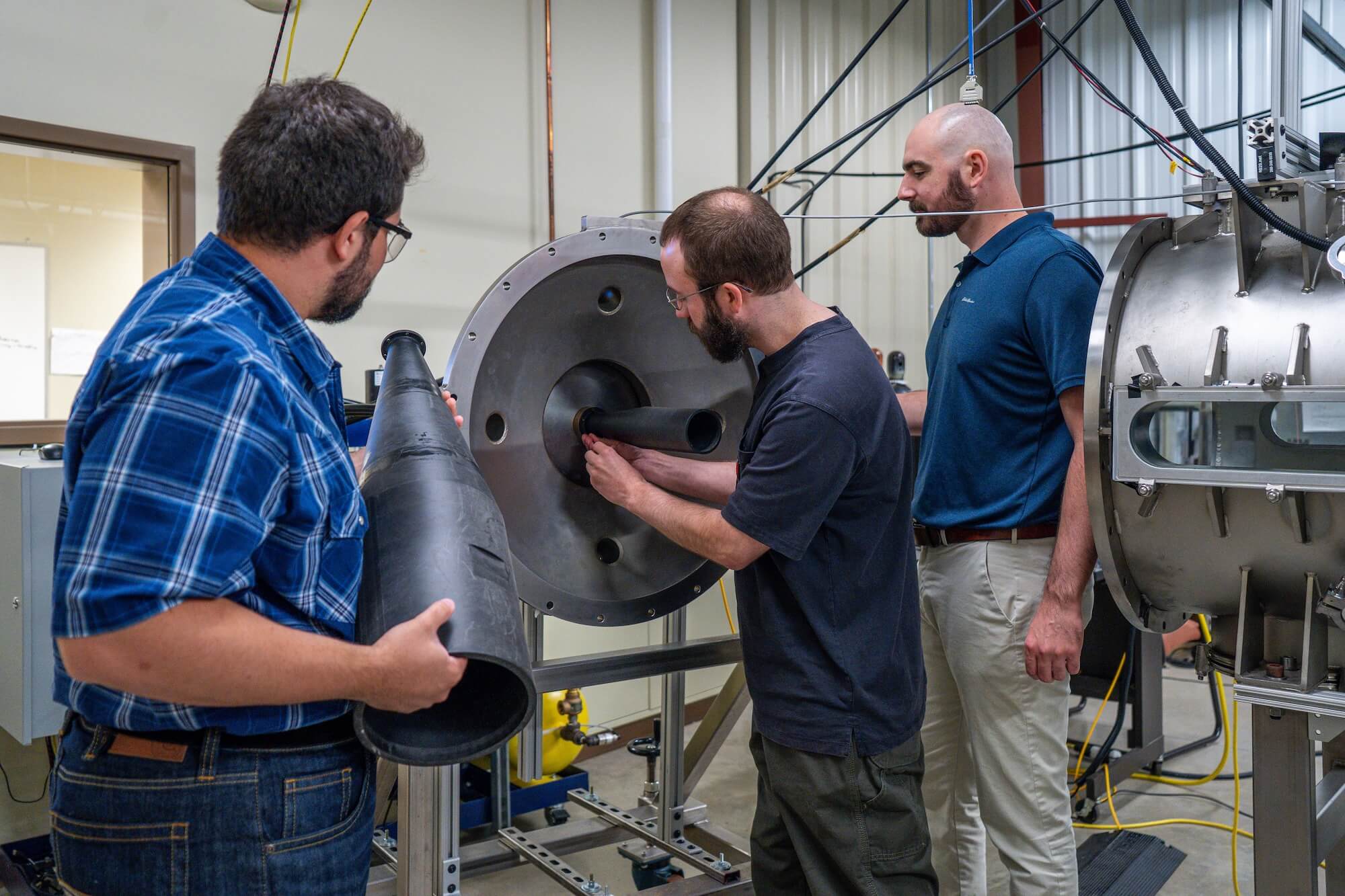 Three men work on scientific equipment in a lab. A man in a blue plaid shirt holds a black conical object, while another with a beard inserts a black tube into a large silver metallic apparatus. A bald man in a blue polo observes the process.