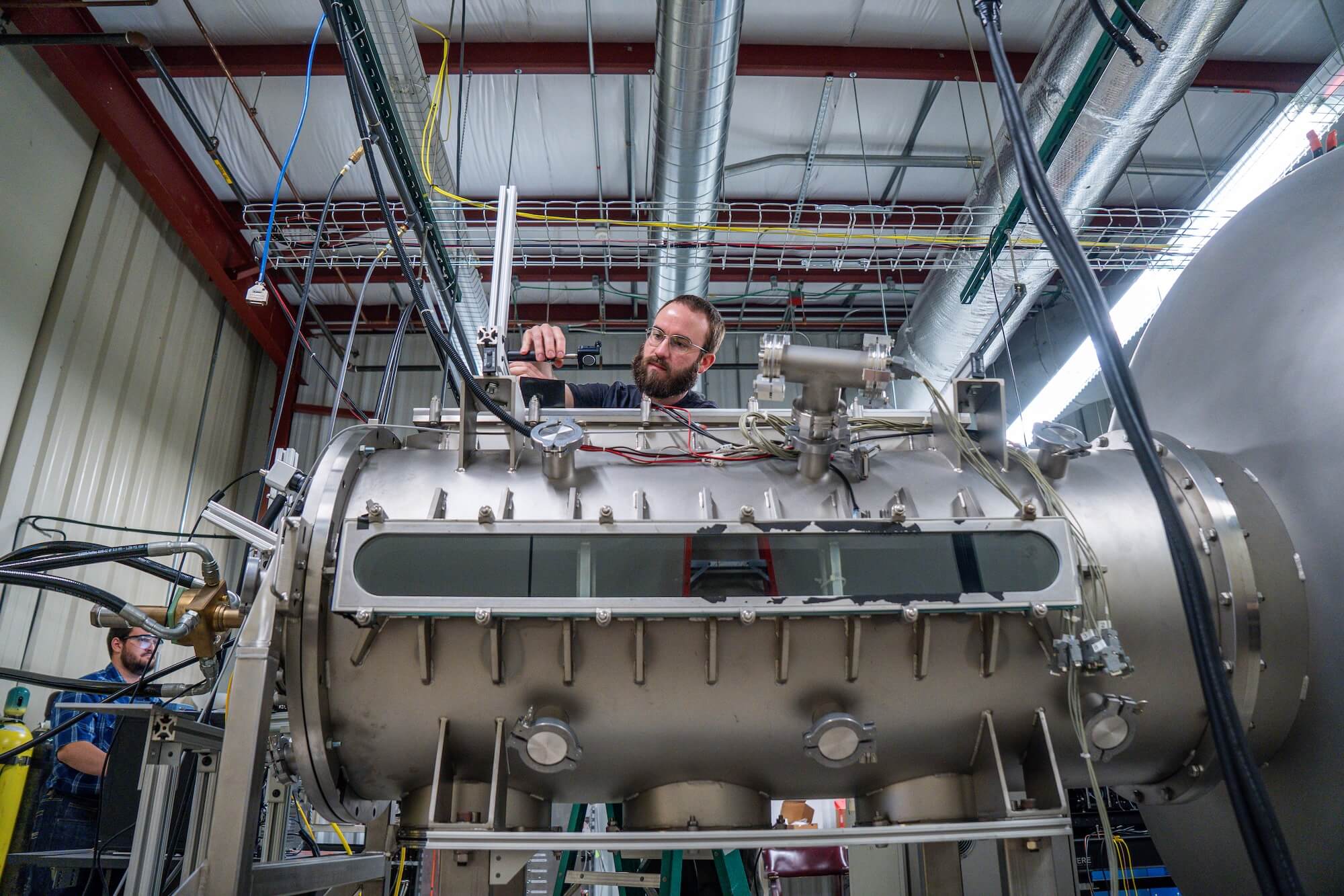 A bearded man in glasses and a black shirt leans over a large, silver cylindrical scientific instrument with a viewing window, carefully adjusting a component with a tool. He is focused in a lab.