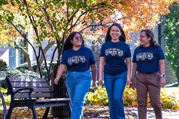 Three smiling women walk right on a Notre Dame campus path. They wear dark blue "Somos ND" shirts amidst colorful fall foliage.