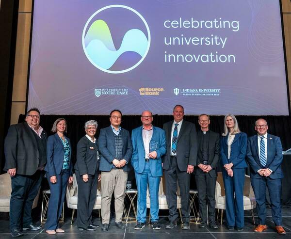 Nine people, including a man holding a crystal award and a man in a clerical collar, smile on a stage. Screen shows "Celebrating University Innovation."