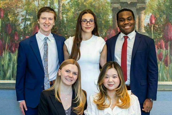 Five smiling students, three standing and two seated, in professional attire against a painting of colorful tulips.