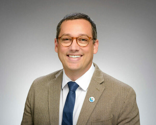 A man with graying hair, tortoise-shell glasses, blue tie, and brown herringbone blazer smiles broadly.