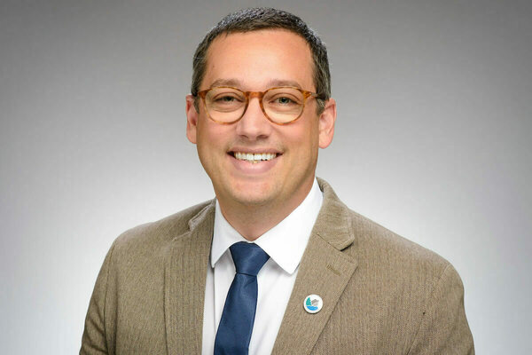 A man with graying hair, tortoise-shell glasses, blue tie, and brown herringbone blazer smiles broadly.