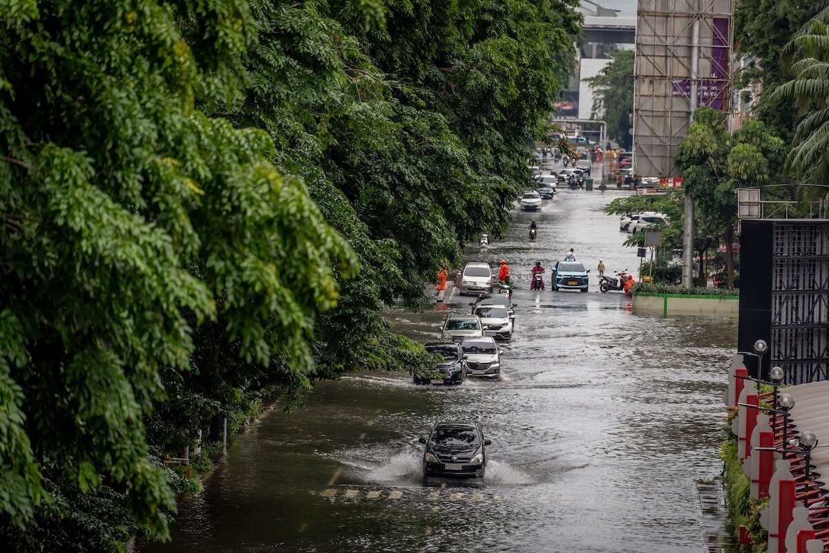 Black and silver cars, motorbikes, and people in orange vests navigate a deeply flooded urban street lined with lush green trees.