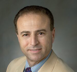Middle-aged man with brown eyes, smiling, in a tan blazer, blue striped shirt, and a dark patterned tie.