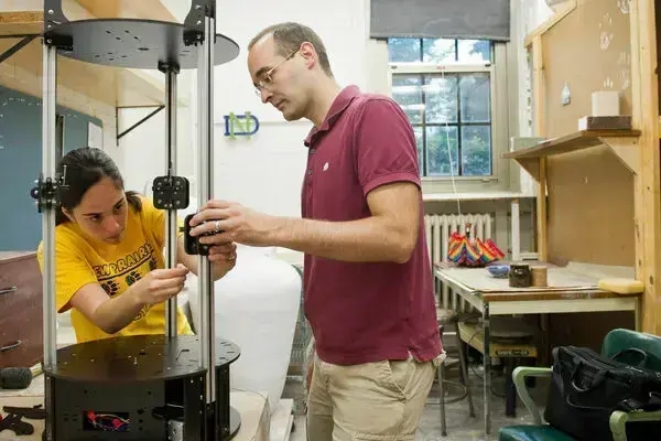 A woman in yellow and a man in maroon intently assemble a tall, black and silver scientific apparatus in a lab.