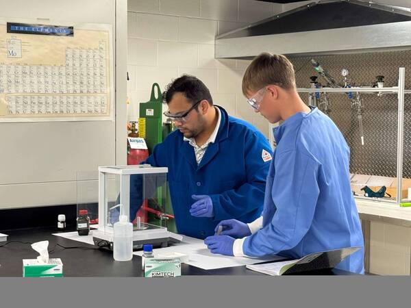 Two researchers in blue lab coats and safety glasses work at a lab bench. One uses a scale, the other looks at notes.