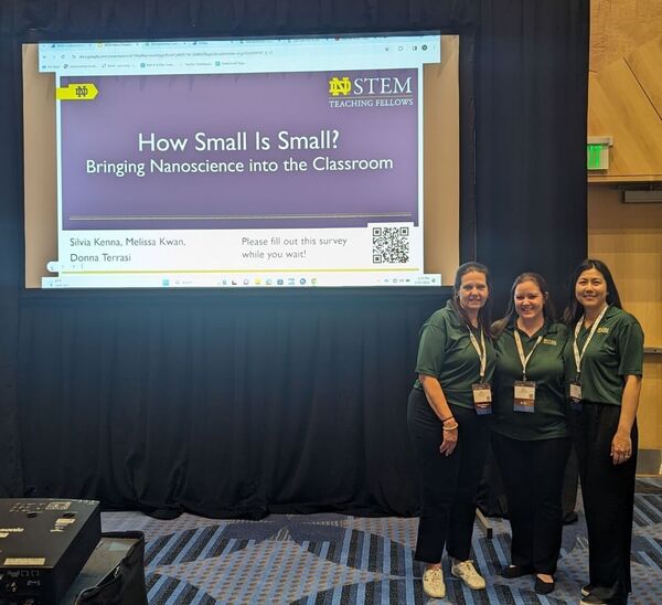 Three smiling women in green ND STEM polos stand by a screen displaying a nanoscience presentation by Kenna, Kwan, Terrasi.