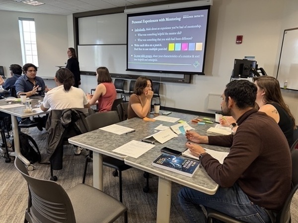 Students in a classroom discuss mentoring experiences around tables. A woman stands by a screen showing a "Personal Experiences with Mentoring" slide.