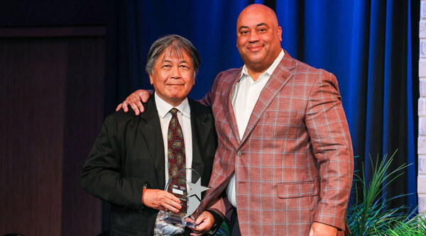 Smiling Asian man with gray hair holds a clear star award. Bald Black man in red plaid jacket puts his arm around his shoulder.