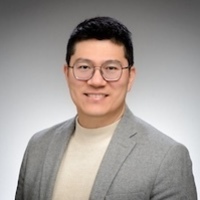 A man with dark hair and glasses smiles. He wears a cream turtleneck sweater under a grey blazer.