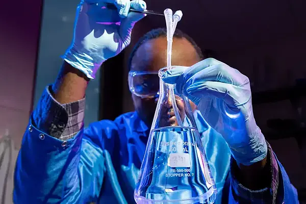 Scientist in blue lab coat and gloves adds liquid to flask using droppers.